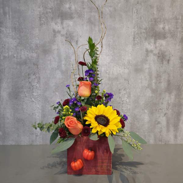 Tall floral arrangement with sunflowers, orange roses, and purple blooms in a red box
