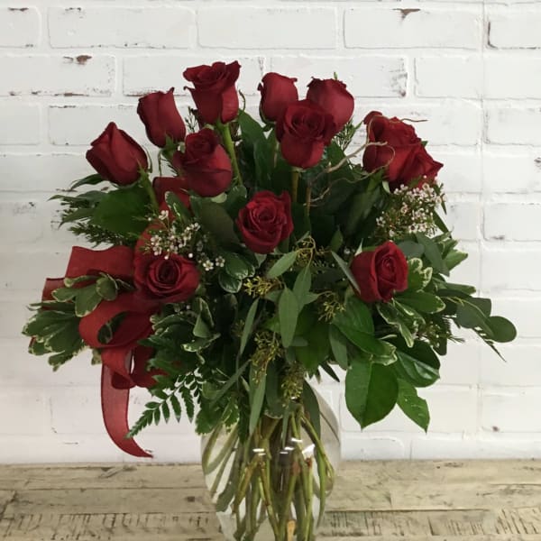 Red roses arranged in a clear glass vase with greenery and a red ribbon