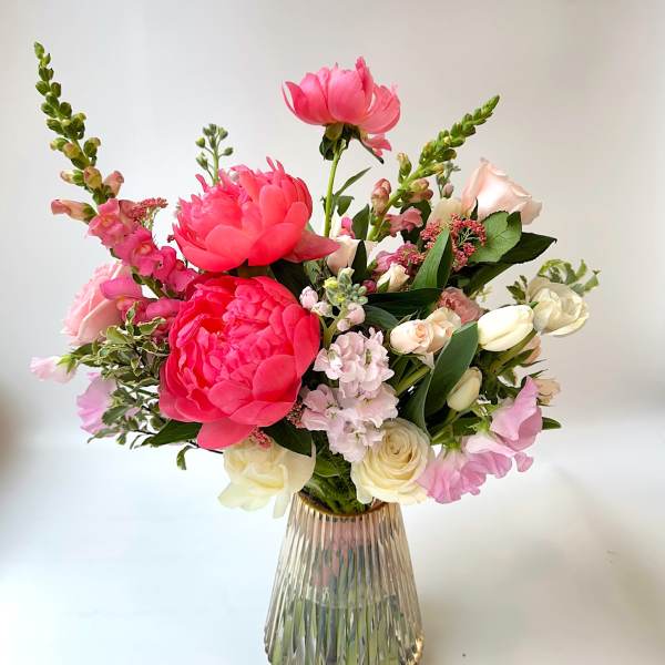 Vase arrangement of bright pink peonies with pale roses, stock, and snapdragons in a ribbed glass vase