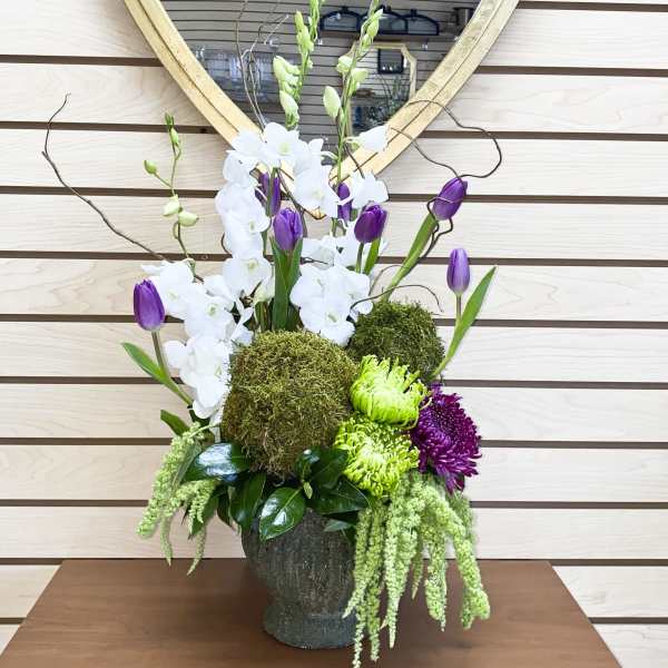 Tall floral arrangement with white orchids, purple tulips, and green moss in a dark vase