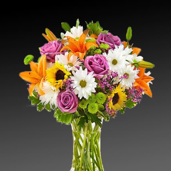 Mixed bouquet of roses, lilies, daisies, and sunflowers in a clear glass vase