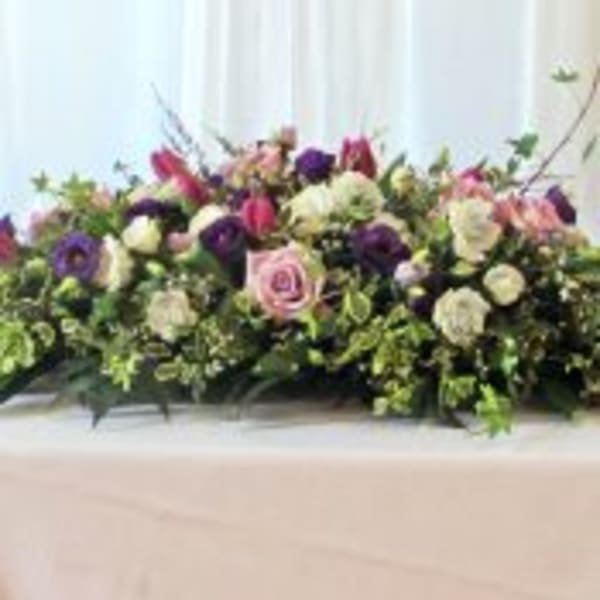Low spray of pink, white, and purple flowers arranged in an elongated display on a draped surface