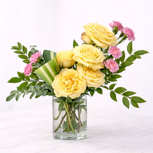 Yellow roses and pink carnations in a clear rectangular glass vase on a light surface