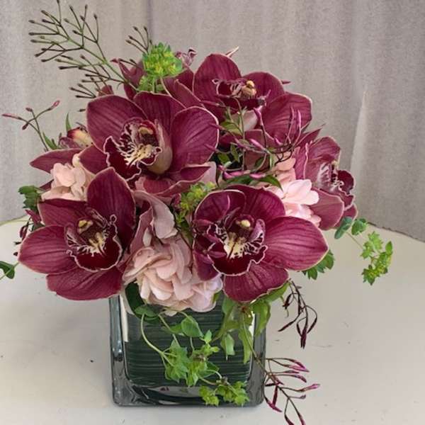 Deep purple orchids arranged in a square glass vase with pale pink blooms.