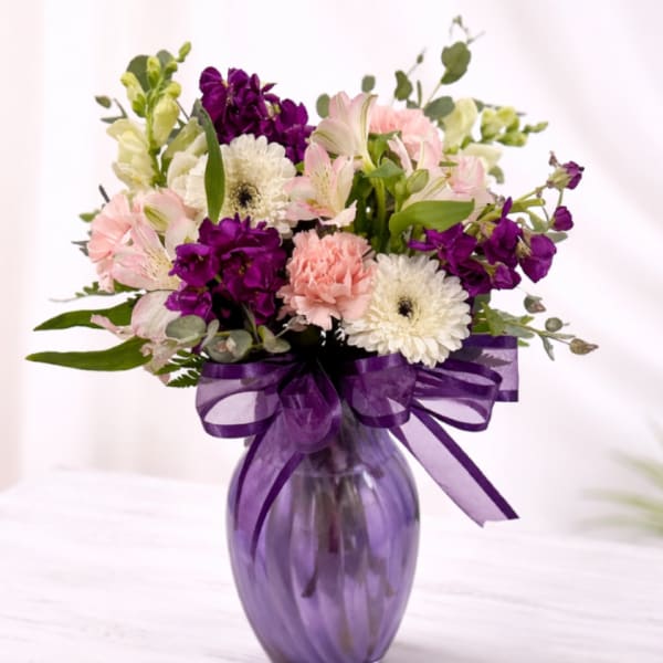Mixed bouquet of pink, white, and purple flowers in a purple glass vase with a matching bow