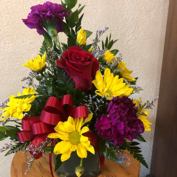 Red rose and yellow daisy arrangement with purple carnations in a glass vase