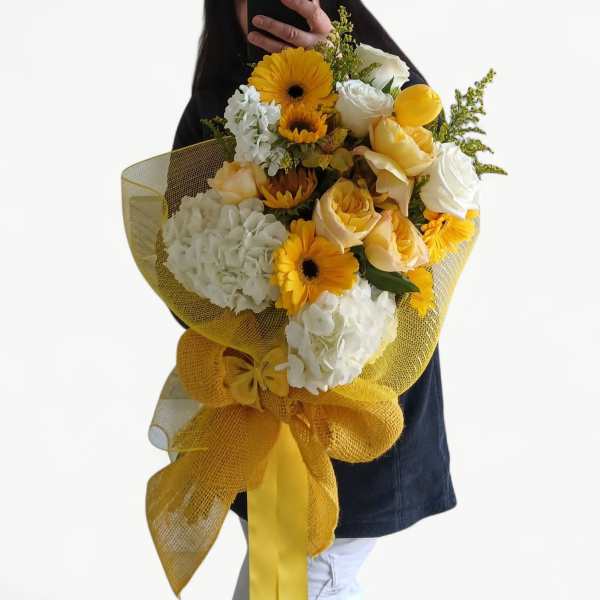 Large hand-tied bouquet of yellow gerberas, white hydrangeas, and roses wrapped in yellow mesh with a bow