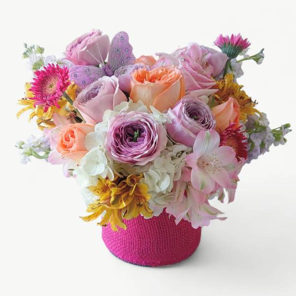 Round arrangement of pink, peach, and yellow flowers in a bright pink fabric-wrapped container with a butterfly pick.