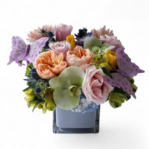 Pastel bouquet of roses, orchids, and mixed flowers in a cube vase with purple butterfly accents
