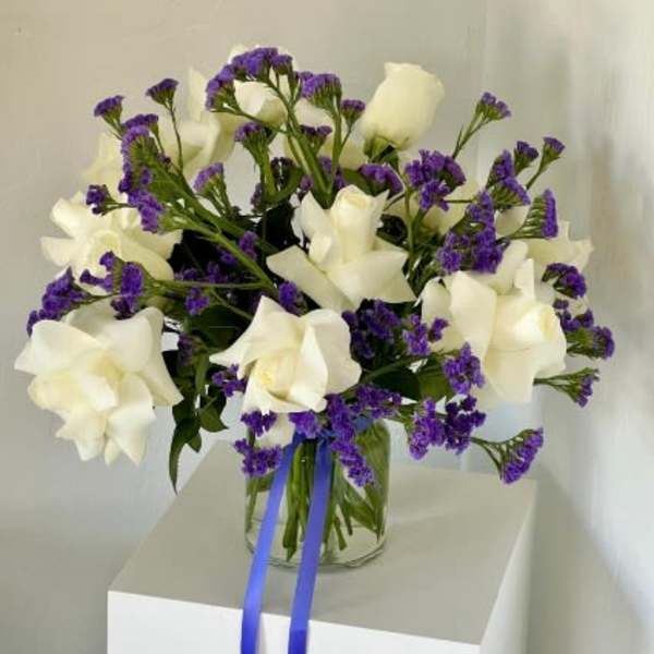 White roses and purple filler flowers in a clear glass vase with blue ribbon
