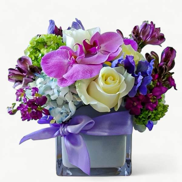 Compact arrangement of pink orchids, white roses, and mixed purple flowers in a cube vase with lavender ribbon