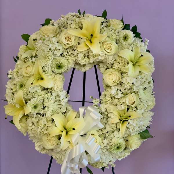Cream floral wreath with lilies, roses, and a white ribbon on a stand