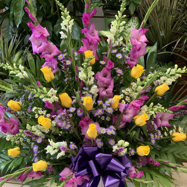 Large mixed floral arrangement with yellow roses, pink gladiolus, and purple daisies in a white vase