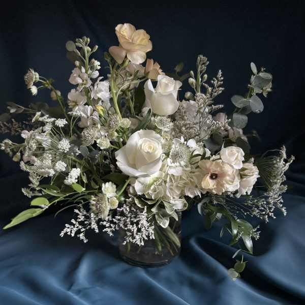 Asymmetrical arrangement of white and cream flowers in a clear glass vase on a dark fabric backdrop