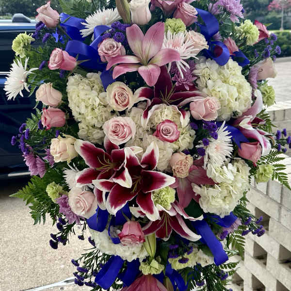Standing spray of pink lilies and roses with white hydrangeas and blue ribbon on an easel