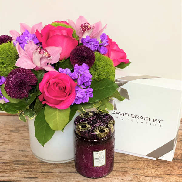 Pink and purple flower arrangement in a white vase with a candle and chocolate box