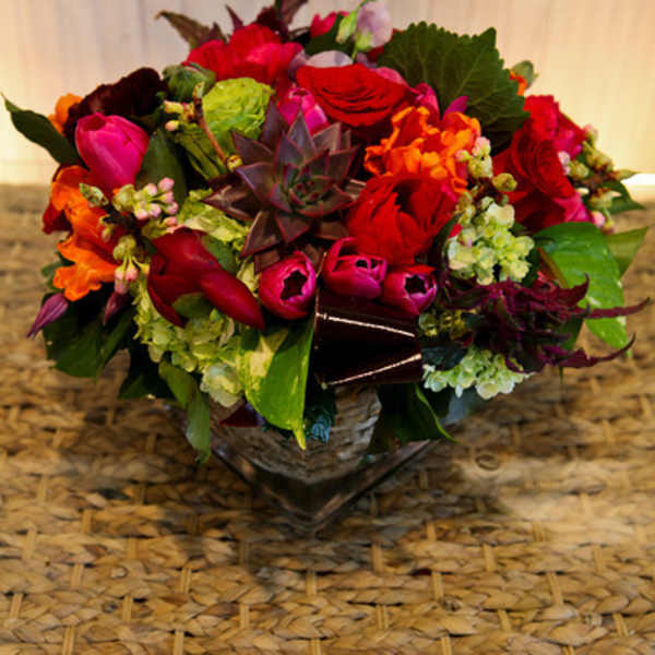 Low mixed arrangement of red roses, pink tulips, green hydrangeas, and a central succulent in a glass cube vase.