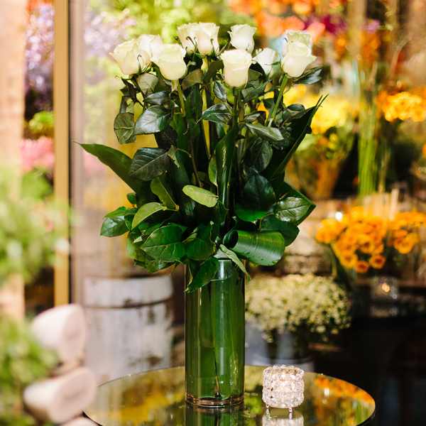 Tall arrangement of white roses with lush foliage in a green glass vase on a table