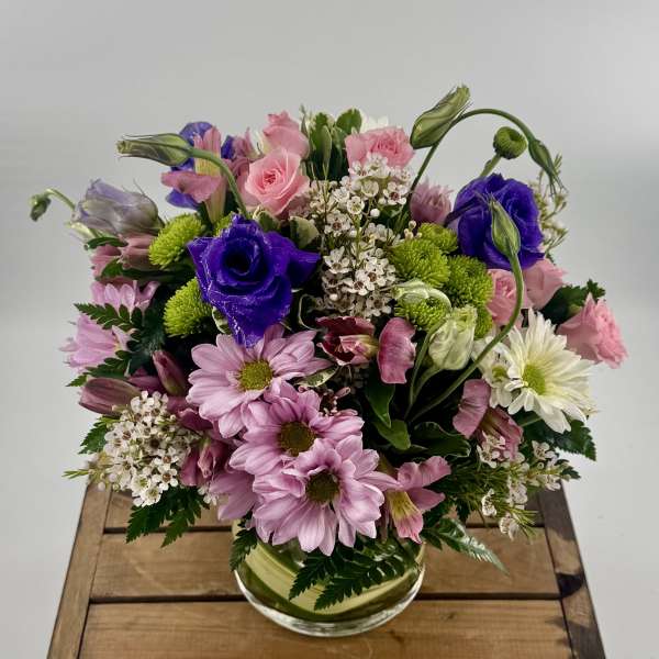 Round arrangement of pink roses, purple lisianthus, and pastel mums in a clear glass vase