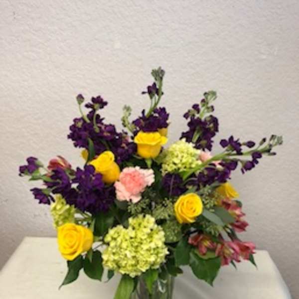 Mixed bouquet in a clear glass vase with yellow roses, purple blooms, and pink carnations
