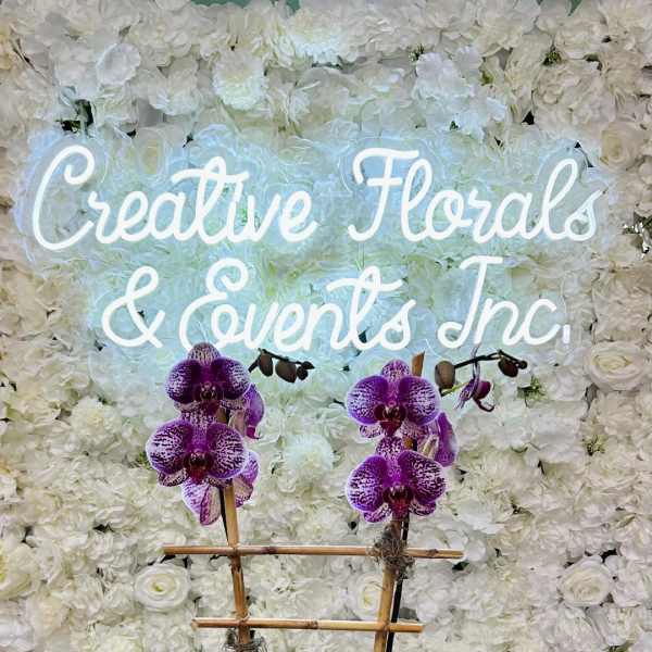 Purple orchids in a black planter before a white floral wall with a neon sign