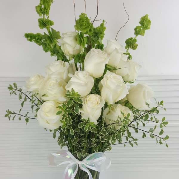 Tall arrangement of white roses and green Bells of Ireland in a clear vase with a white ribbon bow