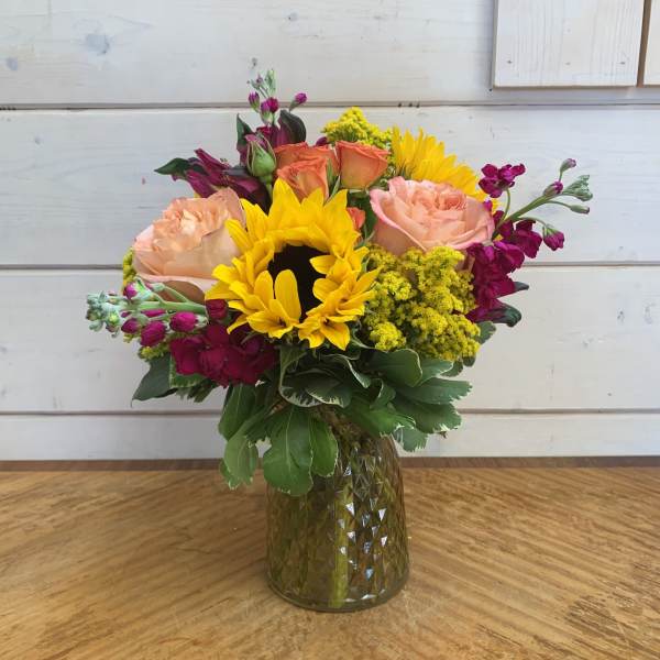 Mixed bouquet with sunflowers, peach roses, and magenta flowers in a glass vase
