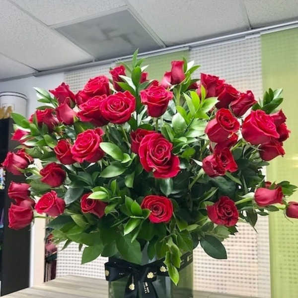 Large arrangement of red roses with abundant greenery in a clear glass cube vase tied with a black ribbon.