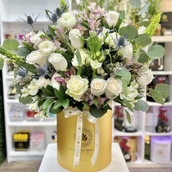 Tall arrangement of white roses and mixed flowers in a gold hatbox-style container with ribbon bow.