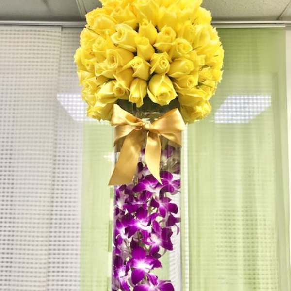 Tall glass vase with submerged purple orchids and a dome of yellow roses tied with a satin bow.