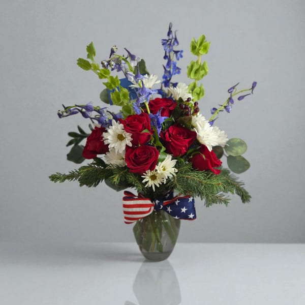 Red roses and white daisies in a glass vase with a patriotic ribbon