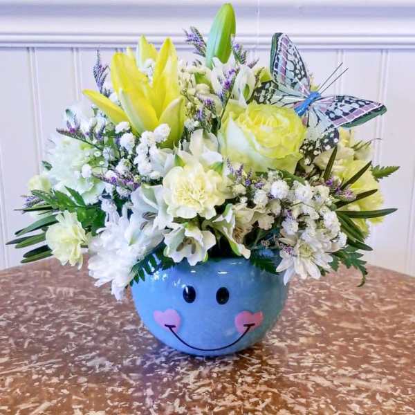 Yellow and white flower arrangement in a blue smiley-face bowl with a butterfly decoration