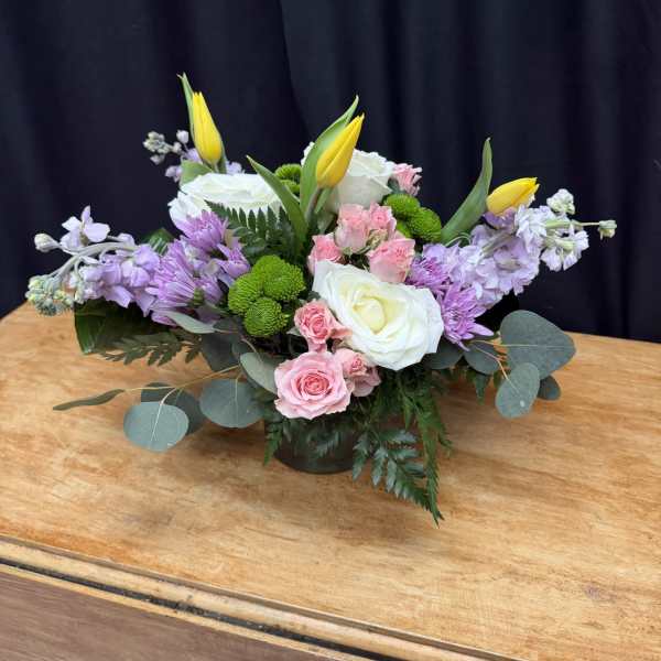 Low floral arrangement with white roses, pink spray roses, purple blooms, and yellow tulips in a vase
