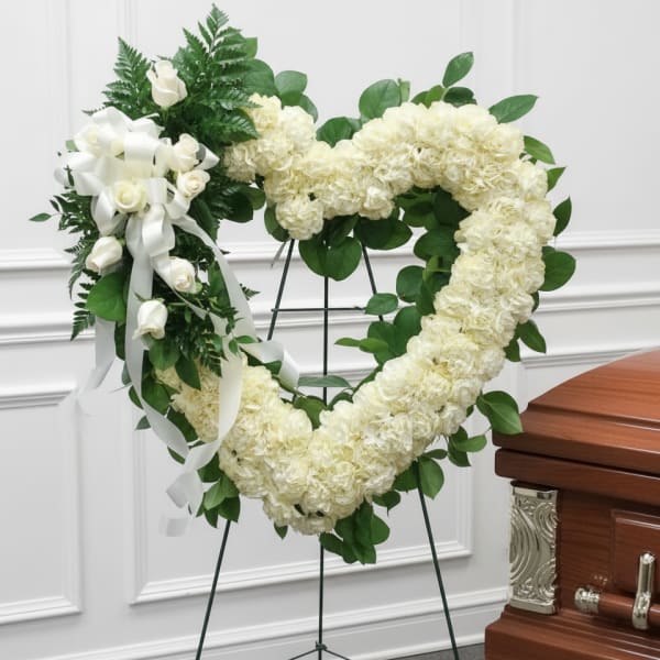 Heart-shaped white floral funeral wreath on a stand beside a casket
