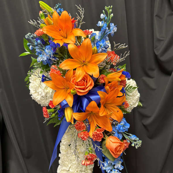 Tall cross-shaped spray of white carnations with orange lilies, roses, and blue flowers on a stand