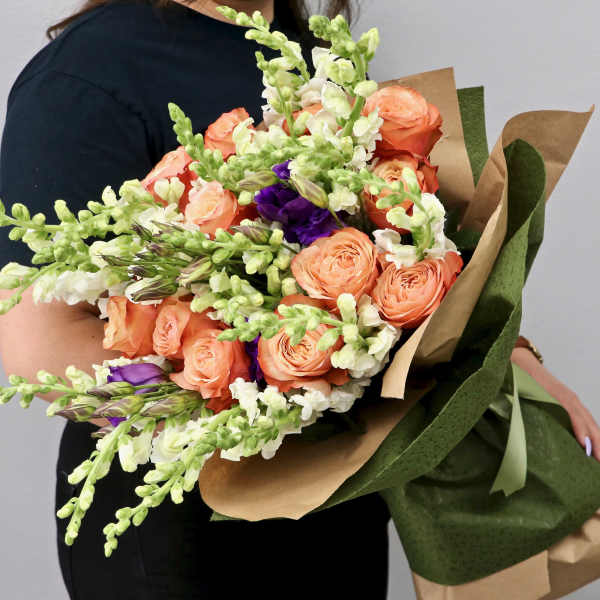 Large hand-tied bouquet of peach roses, white snapdragons, and purple flowers wrapped in kraft paper.