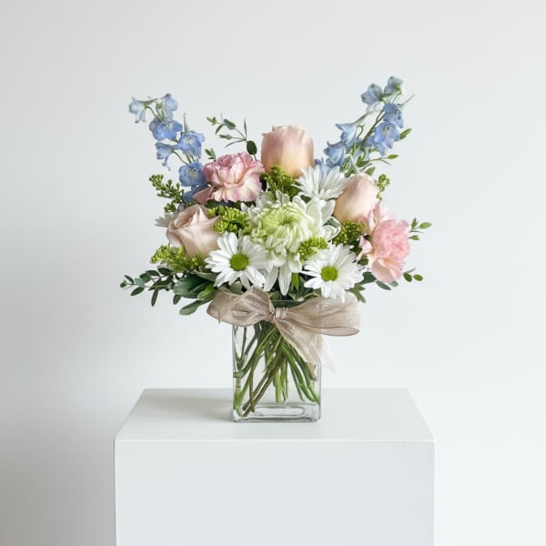 Pastel bouquet of roses, daisies, carnations, and delphinium in a clear glass cube vase with a ribbon bow.