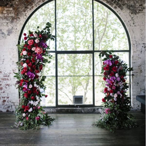 Two tall floral pillars with red, pink, and purple blooms by a large window
