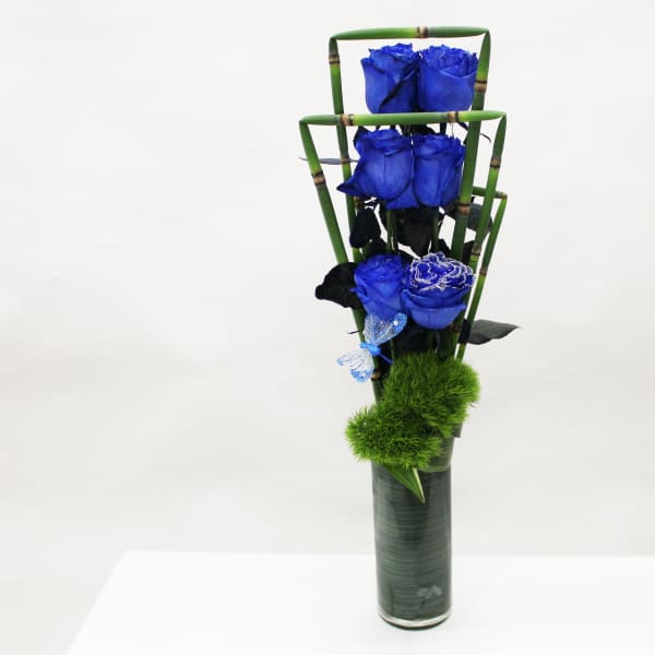 Tall modern arrangement of vivid blue roses in a green-lined glass cylinder with bamboo accents and fuzzy green dianthus
