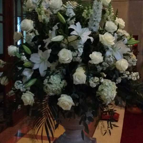 Tall white floral arrangement with roses, lilies, and hydrangeas in a stone urn