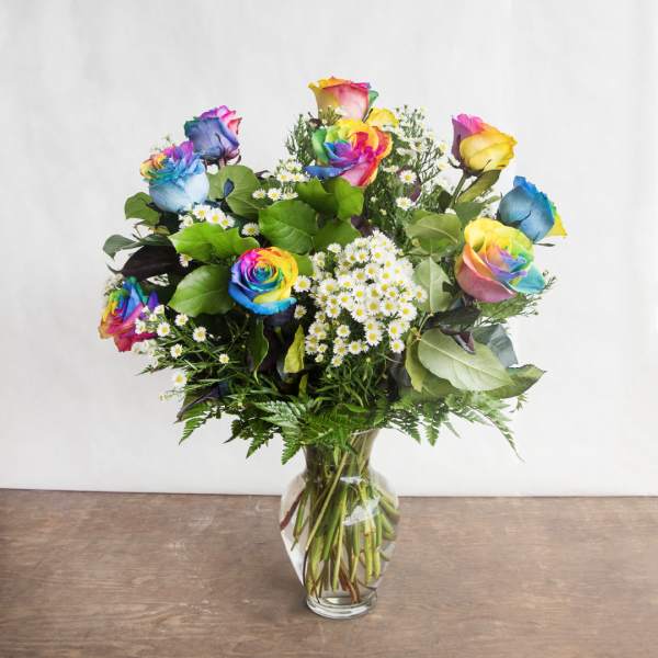 Arrangement of rainbow-colored roses and small white daisies in a clear glass vase on a wooden surface.
