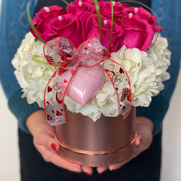 Pink roses and white flowers arranged in a round gift box with a ribbon and heart ornament.
