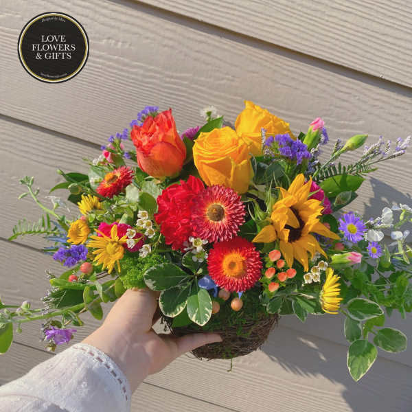 Bright mixed arrangement with yellow roses, red gerberas, and a sunflower in a low twig basket.