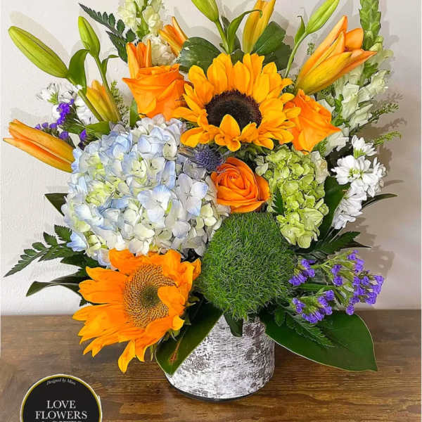 Bright arrangement of sunflowers, orange roses, lilies and hydrangeas in a white cylinder vase