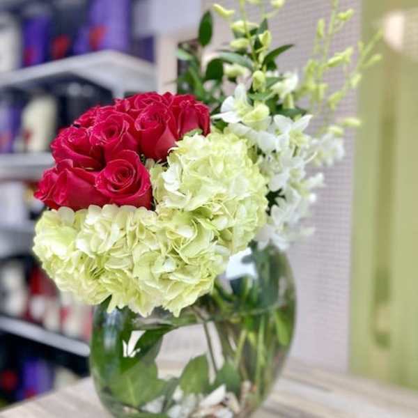 Asymmetrical arrangement of red roses, green hydrangeas, and white orchids in a round glass vase with shells.