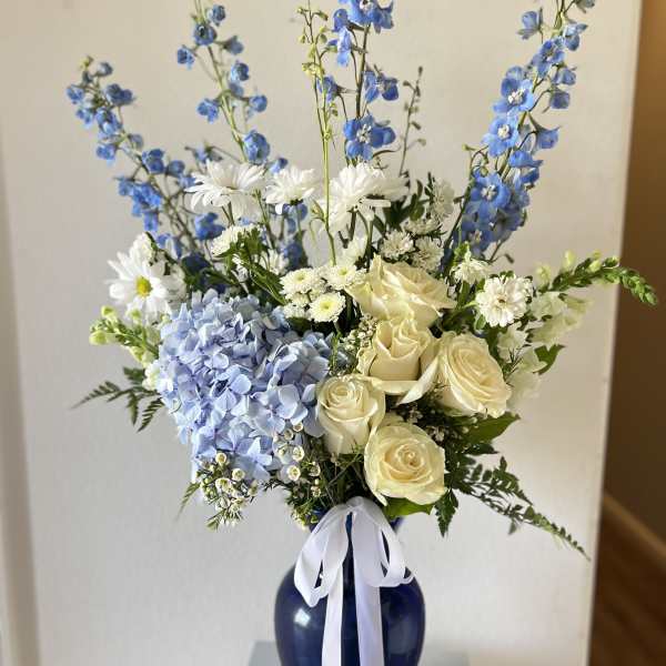 Tall blue and white arrangement with roses, hydrangea, and delphinium in a navy vase with white ribbon.