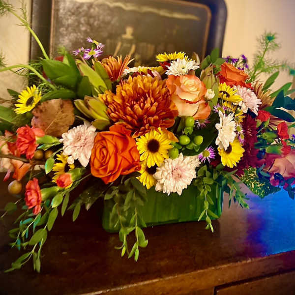 Low centerpiece of orange roses with yellow and white mums in a green rectangular vase