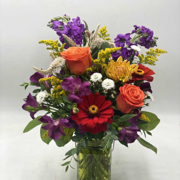 Mixed bouquet of orange roses, purple flowers, and red blooms in a glass vase