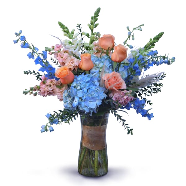 Tall vase arrangement with blue hydrangeas, peach roses, and blue delphinium in a clear glass vase.