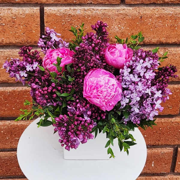 Low arrangement of bright pink peonies and purple lilacs in a white square vase.